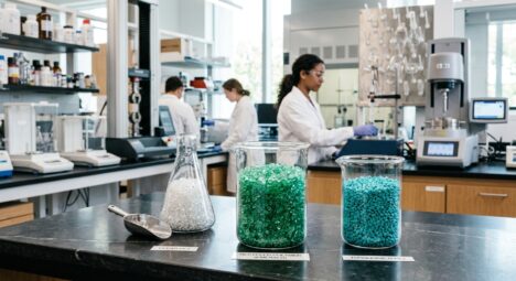 A professional editorial photograph representing modern plastic recycling and material science. In the foreground, vibrant translucent emerald, turquoise, and clear recycled plastic polymer granules sit inside pristine glass laboratory vessels on a sleek studio surface. The background features a brightly lit, modern materials testing laboratory softly out of focus, with clean scientific glassware and subtle high-tech equipment. Crisp lighting highlights the geometric purity, texture, and light refraction of the recycled plastic pellets. Beautiful, clean, eco-tech visual style with rich textures and subtle cinematic depth of field.